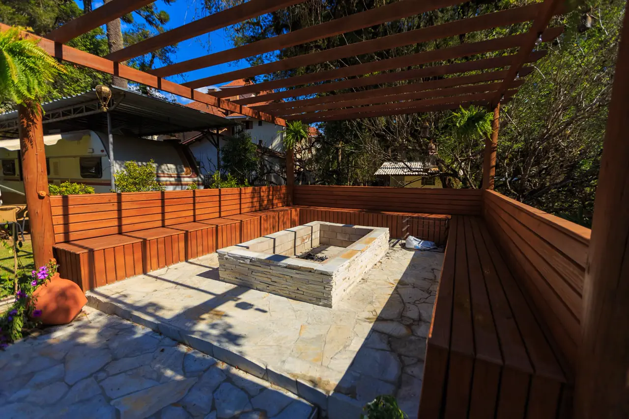 A stone patio with a fire pit and benches under a pergola.